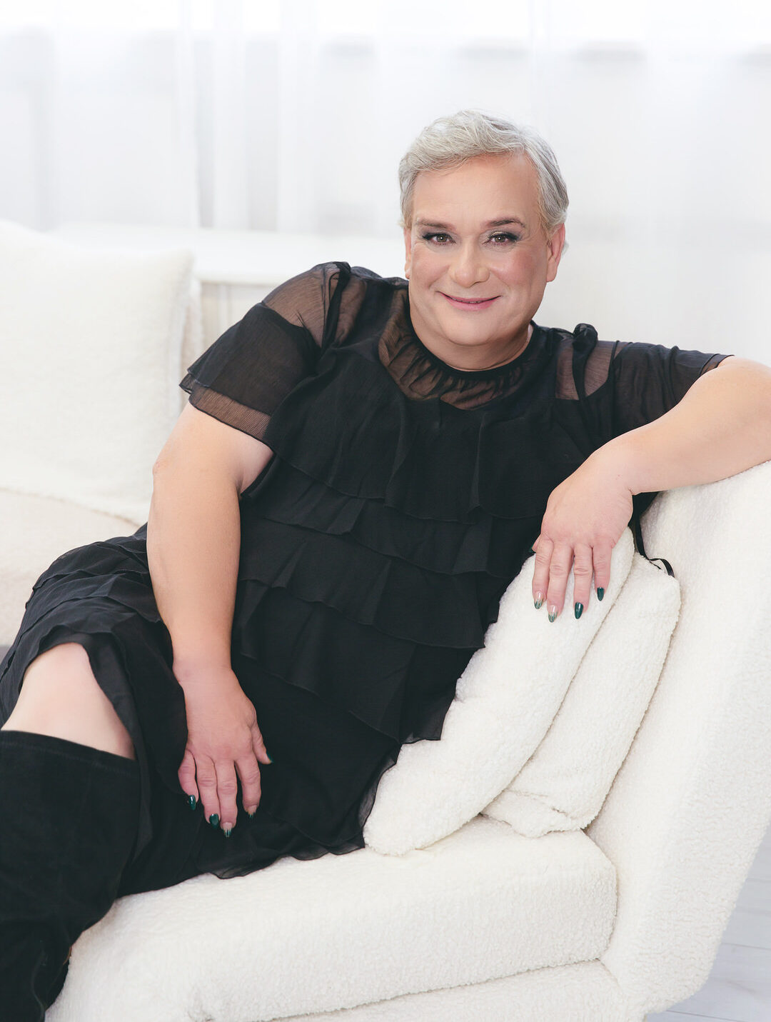 portrait of a cross-dresser with short hair in a black dress and boots sitting on a white sofa