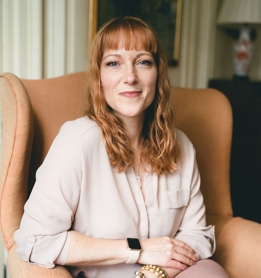 Ginny Marsh sitting in a brown arm chair, wearing a blush coloured shirt and pink pencil skirt