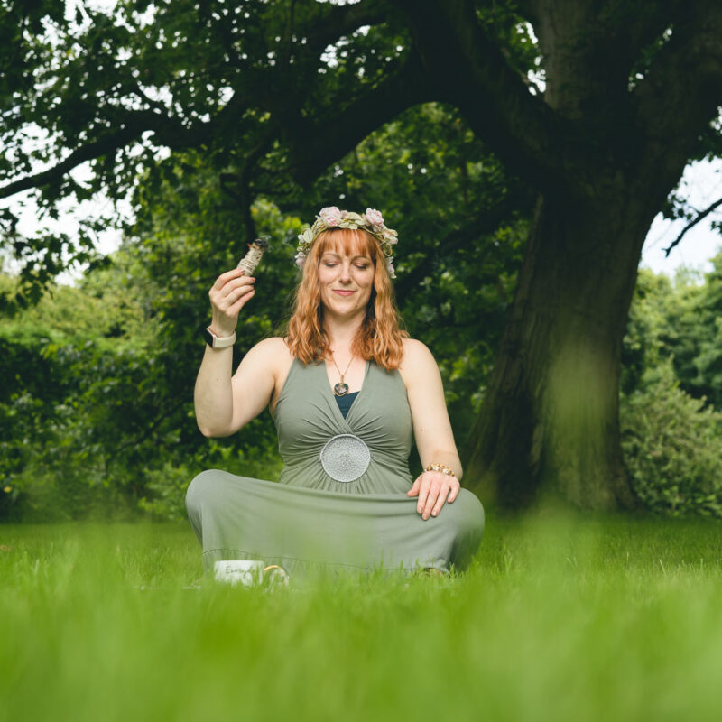 Ginny Marsh wearing a green dress sitting in grass, smudging a space with some white sage